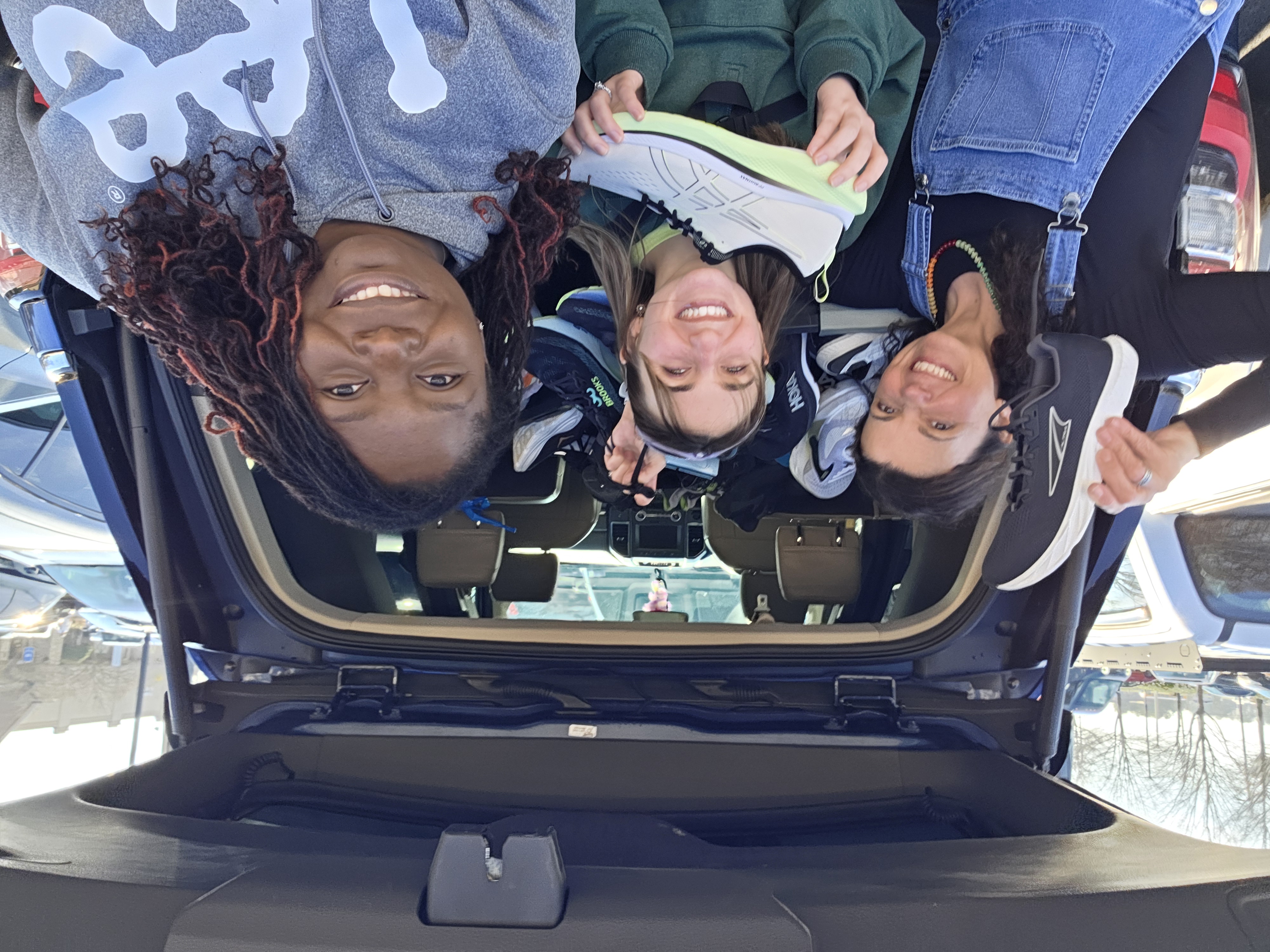 Three women smiling in front of a car trunk full of donated shoes